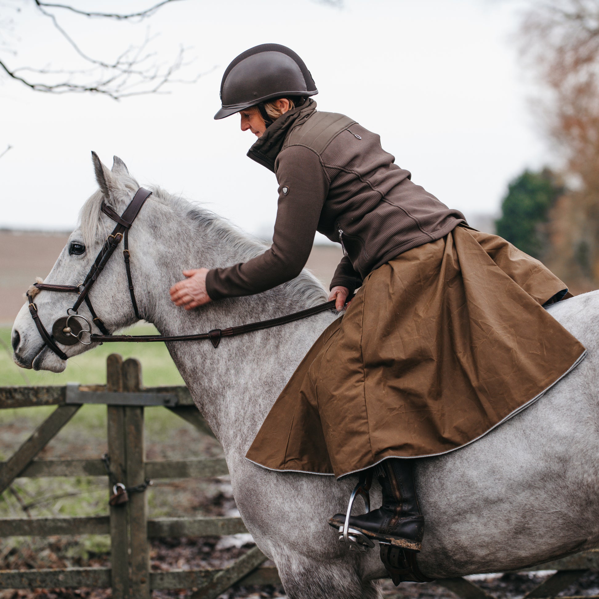 Side view of a woman wearing handcrafted riding habit leaning forward and patting her horse on the neck.