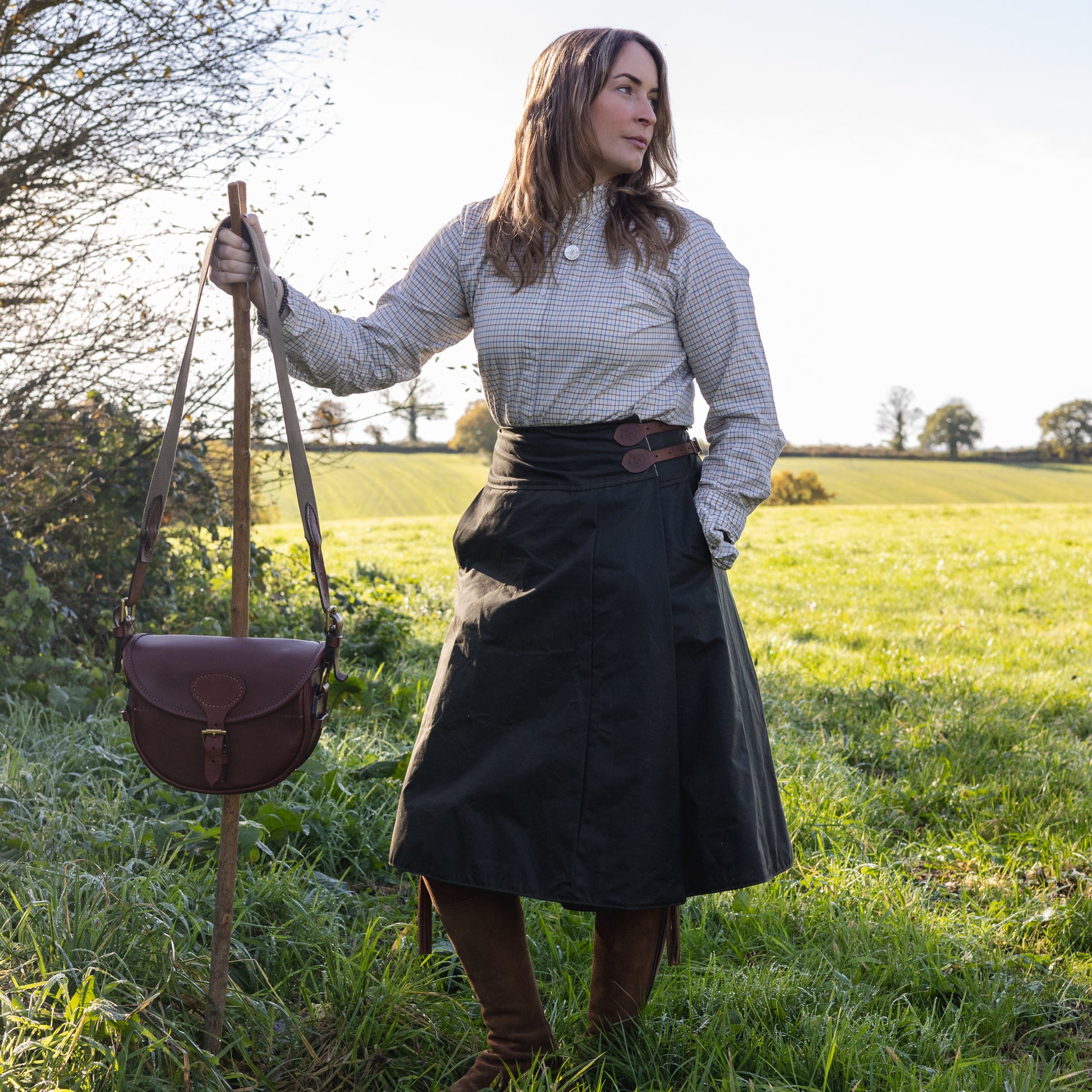 Woman wearing a hand crafted habit stood in a feild with her cartridge case and walking stick ready to go shooting.